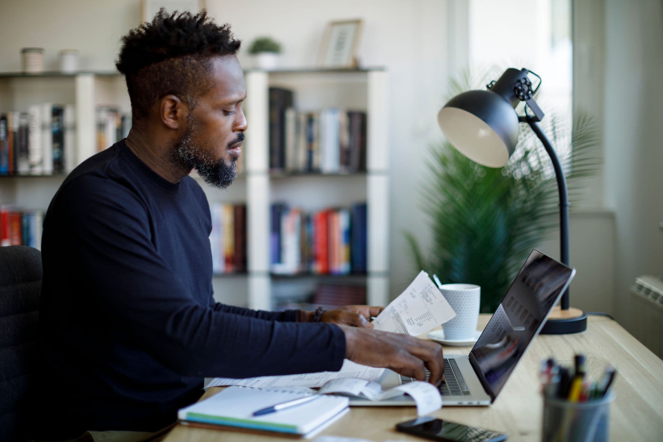 A man sitting at a desk, focused on his laptop, with a notepad and pen beside him.