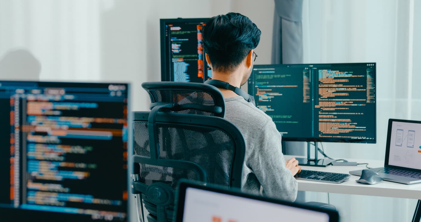 A man working at a desk equipped with two monitors and a laptop, engaged in his tasks.
