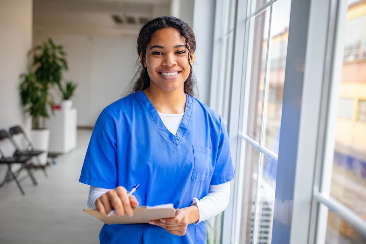 A friendly nurse, holding a clipboard, smiles confidently, embodying compassion and commitment in patient care.