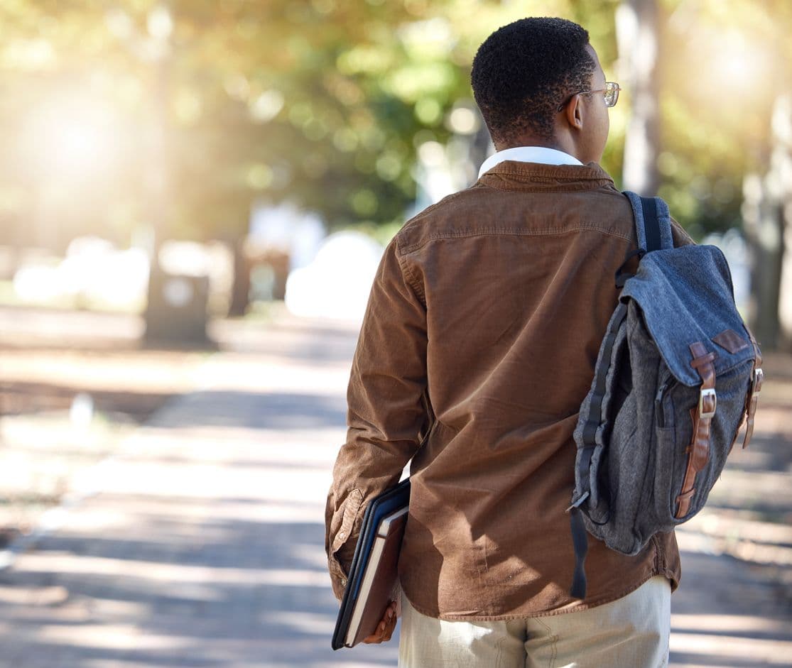 A young man, a college student, walks down a scenic path carrying a backpack, immersed in thought.