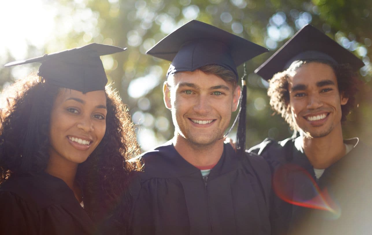 Three joyful graduates stand side by side, smiling at the camera in their caps and gowns, marking their graduation day.