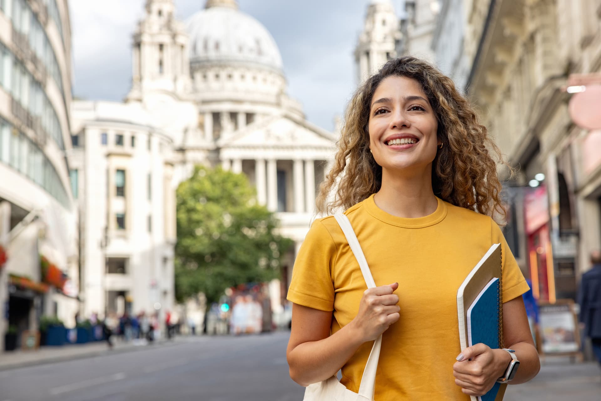 A woman walks on the sidewalk, clutching a book, with a cityscape in the background.